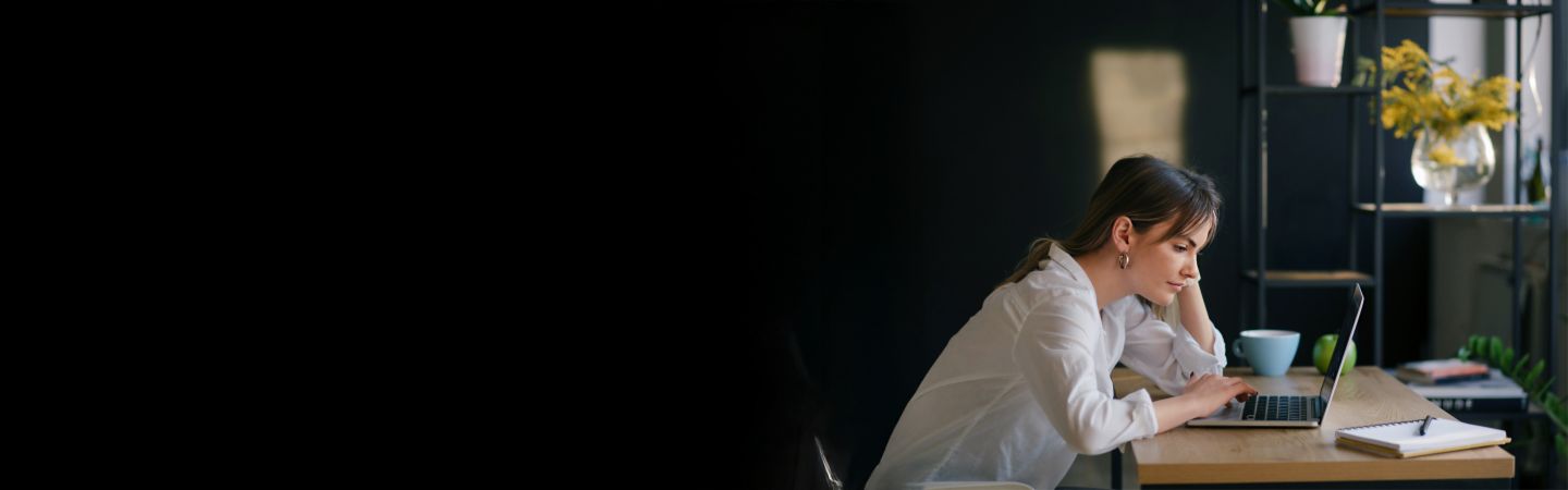 En ung kvinde sidder ved et skrivebord og arbejder koncentreret på en bærbar computer, med en notesbog og en kop ved siden af sig. Baggrunden er mørk, og fokus er på den unge kvinde.<br/>
