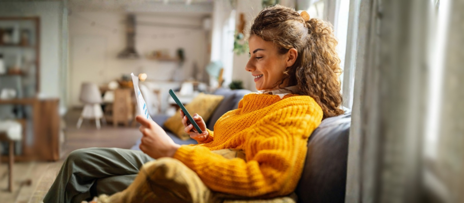 Usmívající se mladá žena s kudrnatými vlasy, v útulném žlutém svetru, sedí na gauči ve svém obývacím pokoji a dívá se na svůj smartphone, zatímco drží papírový dokument.