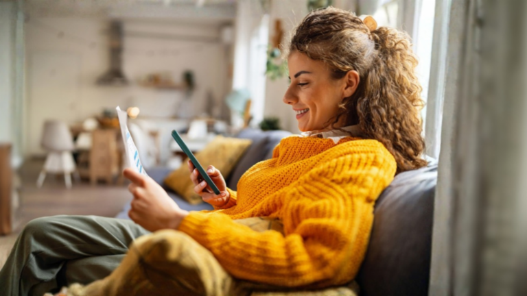 Usmívající se mladá žena s kudrnatými vlasy, v útulném žlutém svetru, sedí na gauči ve svém obývacím pokoji a dívá se na svůj smartphone, zatímco drží papírový dokument.