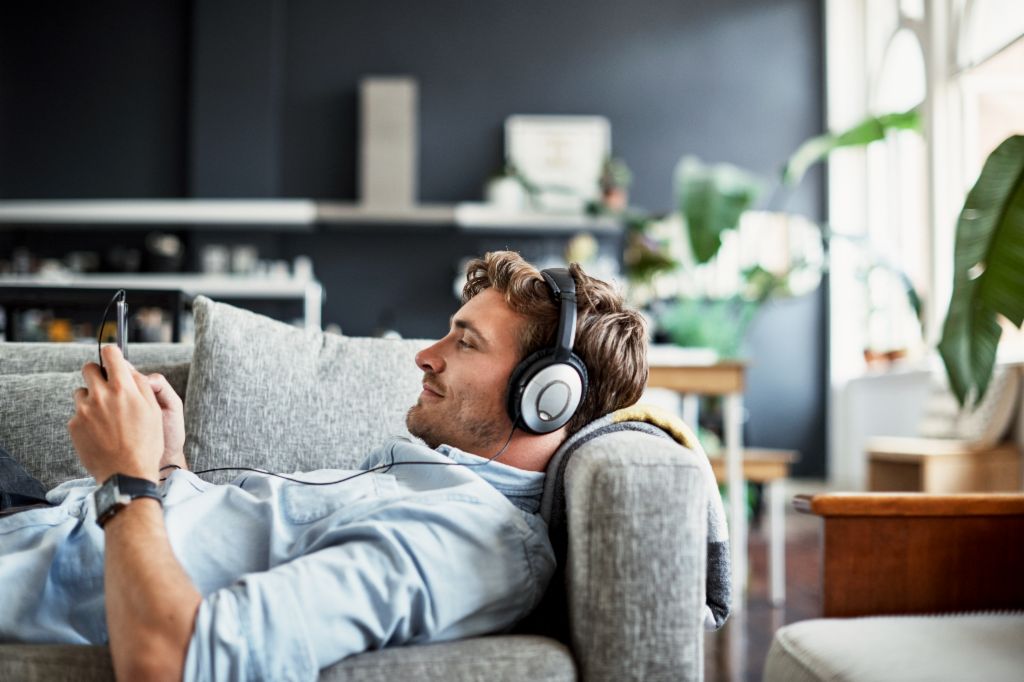 Mladý muž leží na gauči ve svém obývacím pokoji, má na sobě sluchátka a s úsměvem se dívá na svůj smartphone, zatímco si užívá hudbu nebo podcast.