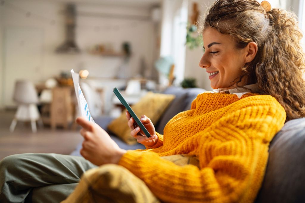 Usmívající se mladá žena s kudrnatými vlasy, v útulném žlutém svetru, sedí na gauči ve svém obývacím pokoji a dívá se na svůj smartphone, zatímco drží papírový dokument.