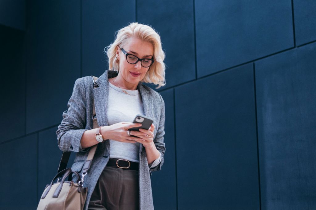 Blond podnikatelka v šedém saku stojí před moderní budovou a usmívá se při pohledu na svůj smartphone.