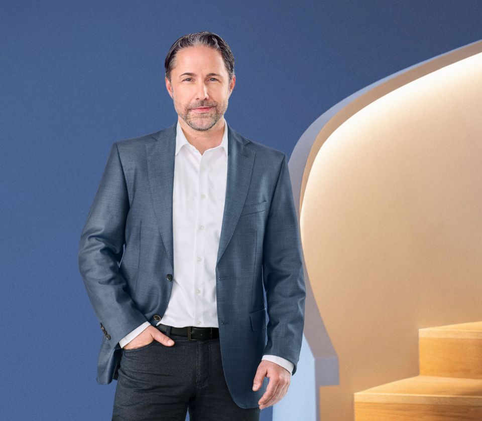 Marwin Ramcke, CEO skupiny EOS, stojí před modrým pozadím, oblečený v tmavém saku. Dívá se přímo do kamery.