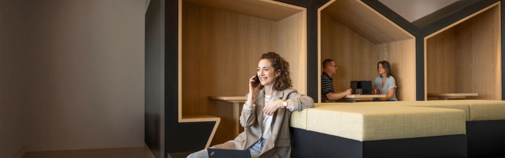 Usmívající se osoba sedí v moderním kancelářském prostředí, telefonuje a pracuje na notebooku. V pozadí pracují další dva lidé.