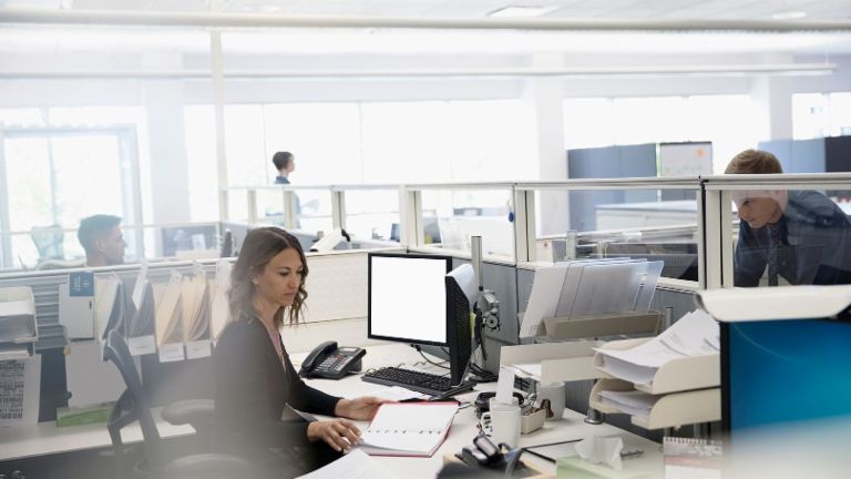 Digitalizace: Tři zaměstnanci v open space kanceláři, jedna žena sedí mezi papíry a svým počítačem