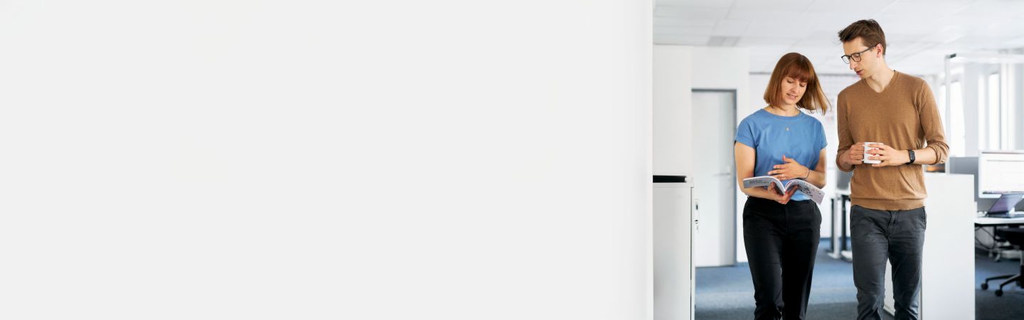 Dva kolegové diskutují v moderní kanceláři o tom, jak může EOS chránit společnosti před platebními výpadky a maximalizovat jejich likviditu v řízení pohledávek, zatímco další zaměstnanec pracuje na počítači v pozadí.