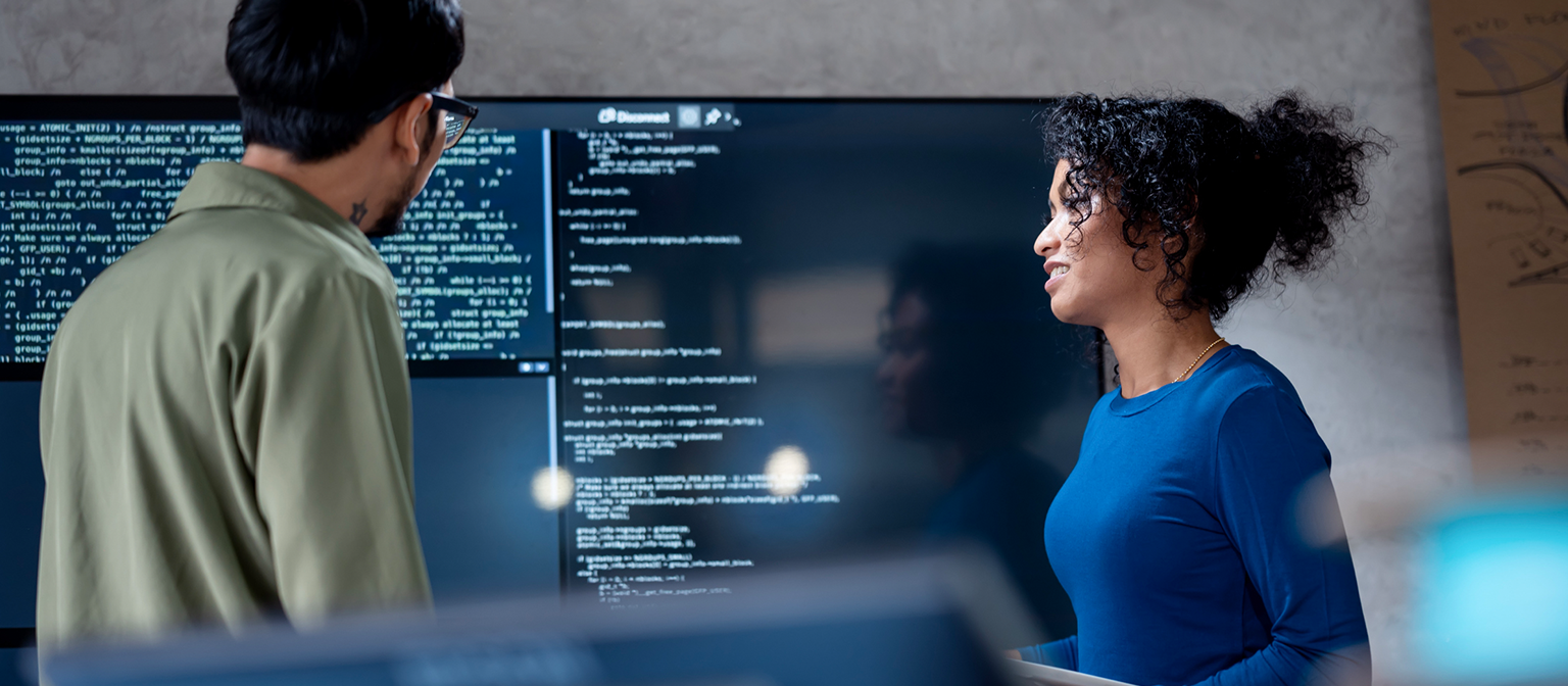 AI v inkasu: Na tomto obrázku dva profesionálové společně analyzují programový kód pro automatizované procesy na velkém displeji. Scéna ilustruje praktickou aplikaci umělé inteligence (AI) a robotizované automatizace procesů (RPA) v řízení pohledávek – příklad digitální transformace v managementu pohledávek.