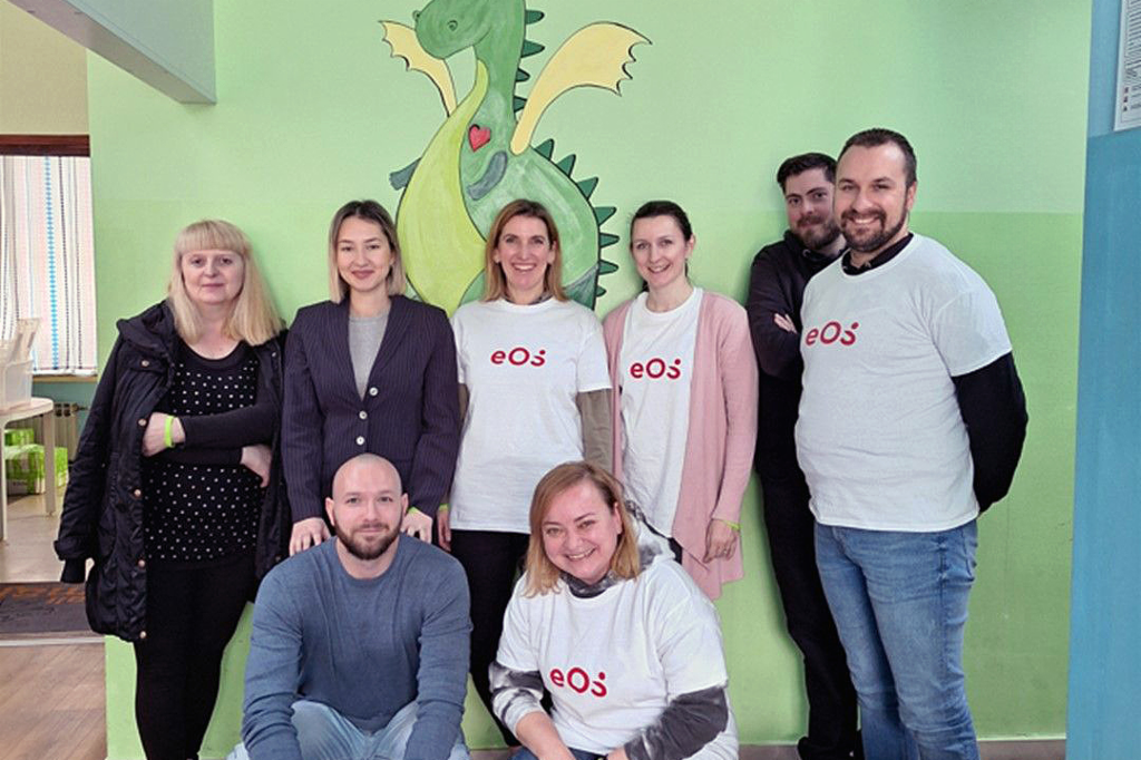 Osm lidí stojí před zdí, na které je namalován zelený drak. Lidé jsou rozmístěni ve dvou řadách: dva dřepí vpředu a šest stojí vzadu. Čtyři lidé mají na sobě bílá trička s červeným logem EOS. Skupina se usmívá do kamery. Na podlaze vpředu vpravo vidíme několik lahví a nápojových kartonů.