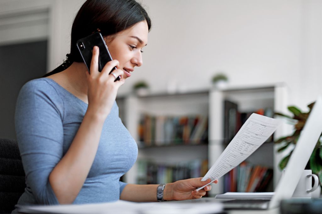 Žena v modrém topu na mobilním telefonu, zatímco drží fakturu v rukou.